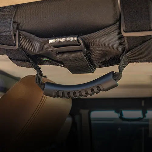 Close-up of a black grab handle with a textured grip attached to the interior roof of a vehicle, near the passenger seat, using nylon straps and a buckle.